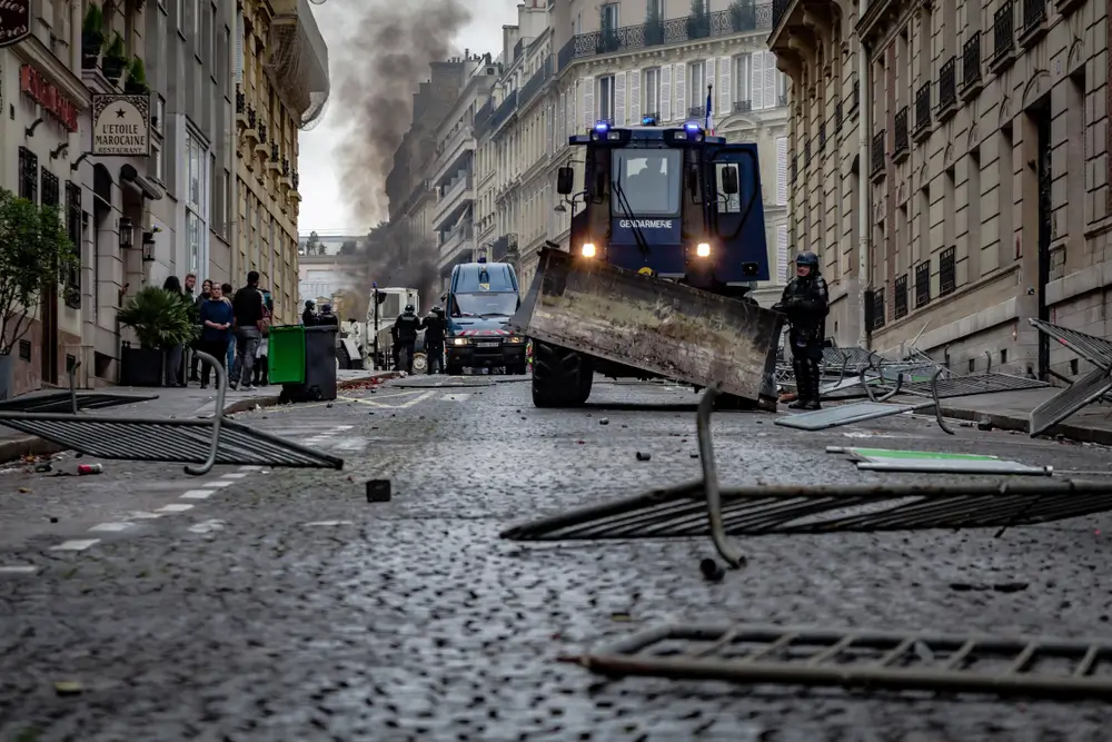Gendarmes en intervention urbaine avec véhicule blindé sur rue pavée, débris au sol et fumée en arrière-plan