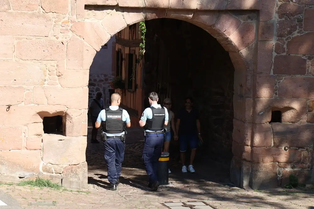 Deux gendarmes en patrouille sous une arche en pierre dans les rues pavées d'un village rural français.