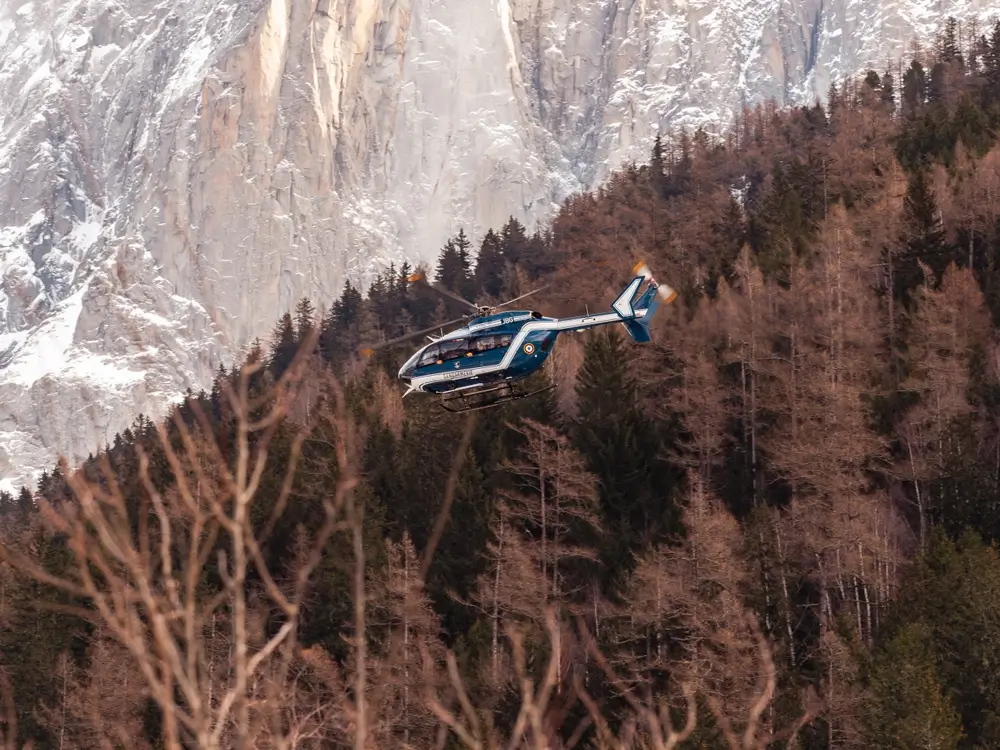 Hélicoptère de la gendarmerie nationale survolant une forêt de montagne devant un massif rocheux enneigé.