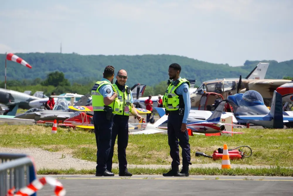 Trois gendarmes de l'air en mission de surveillance sur le tarmac d'une base aérienne devant des avions militaires.