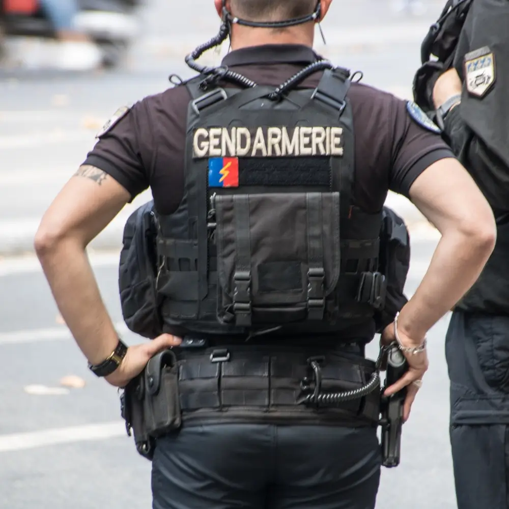 Gendarme de dos en uniforme militaire avec gilet tactique marqué Gendarmerie et ceinture d'équipement réglementaire.
