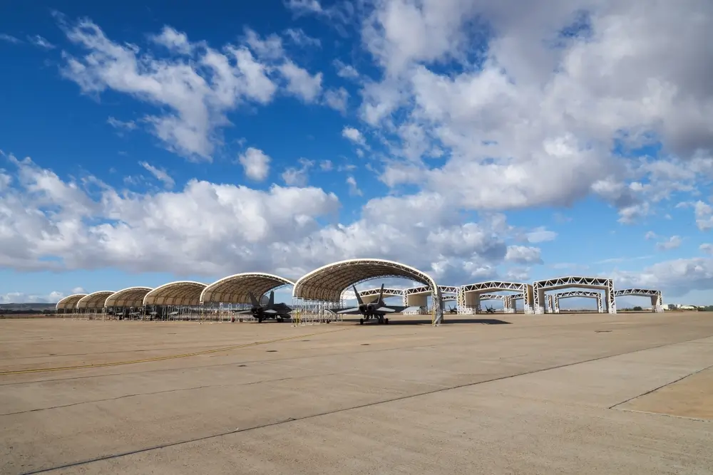 Avions de chasse stationnés sous des hangars sur le tarmac d'une base aérienne militaire, sous un ciel bleu nuageux.