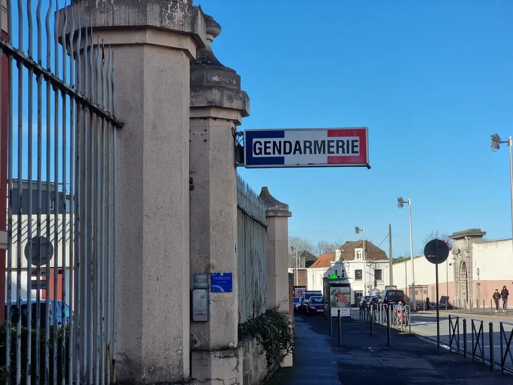 Entrée d'un bâtiment de gendarmerie avec l'enseigne tricolore fixée sur un pilier en pierre bordant une rue.