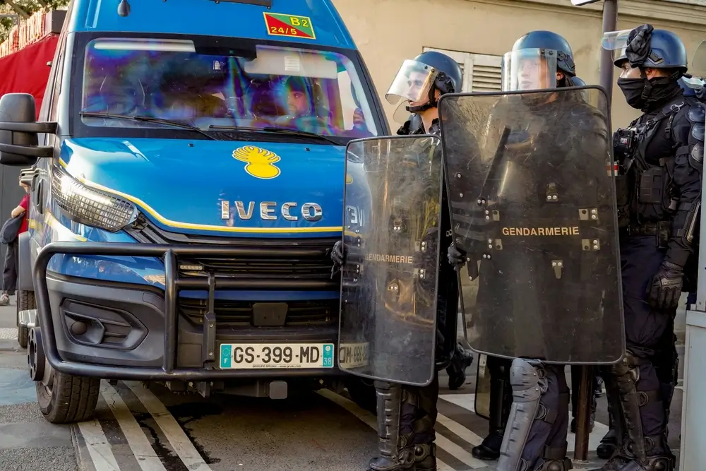 Gendarmes mobiles en tenue de maintien de l'ordre avec boucliers, positionnés devant leur fourgon d'intervention bleu.